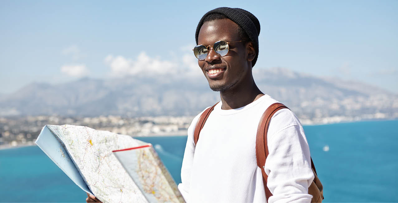 Happy handsome young dark-skinned male traveler standing on top of mountain with paper map above vast ocean and resort town, having joyful look while traveling around the world in company of friends